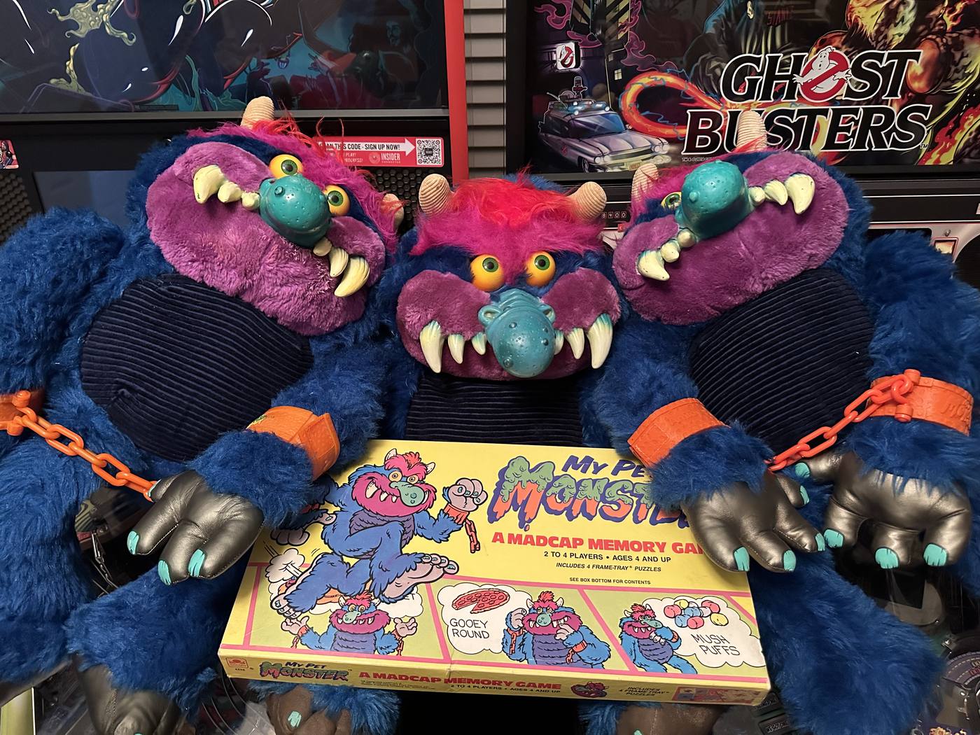 Three vintage My Pet Monster plush toys with orange chain cuffs posed in front of Ghostbusters pinball machines, holding the My Pet Monster Madcap Memory Game